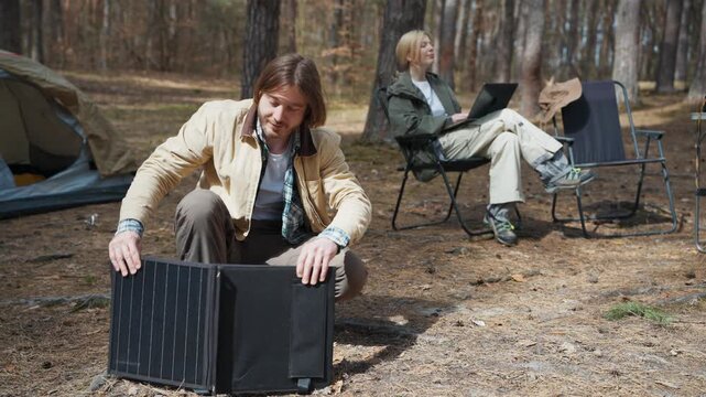 Caucasian man setting portable solar panel near camping tent in forest while blonde woman using laptop outdoors. Hiking couple creating remote workspace with renewable energy during adventure.