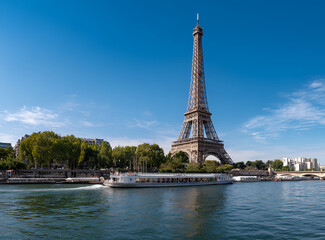 Fototapeta premium A classic view featuring an iconic wrought-iron lattice tower standing tall under a vibrant blue sky with a tourist boat on the water