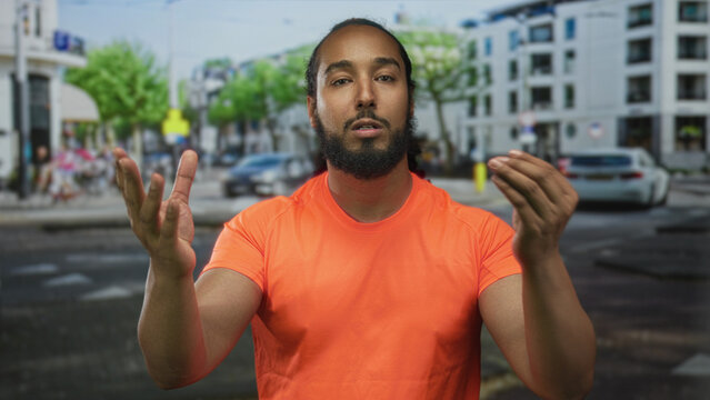 Man with finger to lips for silence and open hands on a city street near buildings wearing orange shirt; contemplation quiet appeal.