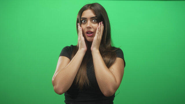 Woman covers mouth with hands, wide eyes and burgundy painted nails facing camera against green studio backdrop; surprise curiosity.