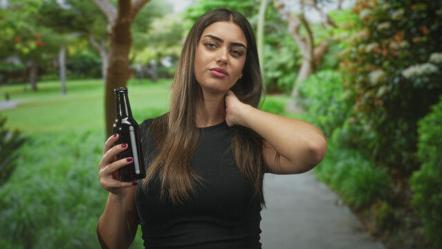 Woman holding dark beer bottle with hand on neck, fingernails painted, standing on a paved path in forest near trees and shrubs; pensive contemplation.