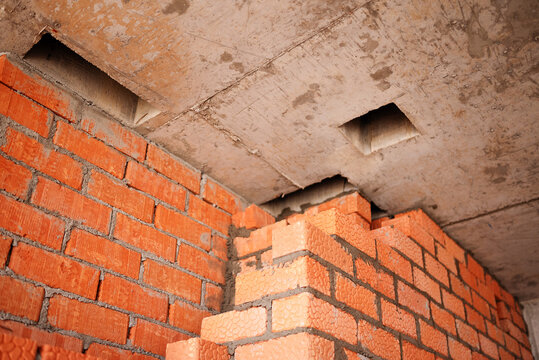 An unfinished interior partition wall made of textured red clay bricks is being built up to meet a rough concrete ceiling featuring square ventilation shafts.