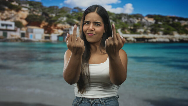 Woman showing middle fingers with both hands, grimacing while wearing white tank top and denim jeans at a sunny beach; defiance rebellion.