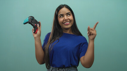 Young woman gamer holding a teal and black gamepad, smiling and pointing index finger upward in studio with teal backdrop and blue shirt  playful confidence. © Krakenimages.com