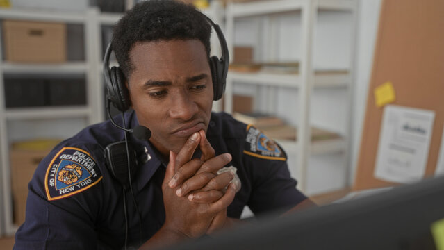 Man police officer with headset, hands clasped at mouth while watching a monitor at building dispatch desk; duty concern.