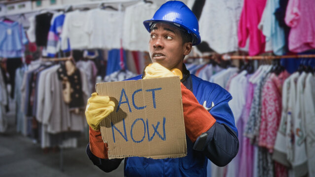 Man builder in hardhat holding cardboard sign reading act now amid clothing racks inside building wearing yellow gloves and blue uniform; urgent solidarity.