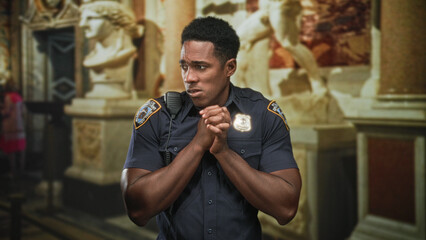 Fototapeta premium Man police officer clasps hands at chest looking upward amid marble statues in ornate building; duty concern.