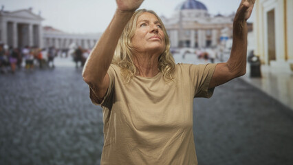 Senior blonde woman raising both arms with bare forearms visible in a busy piazza before a basilica, looking upward  quiet contemplation. © Krakenimages.com