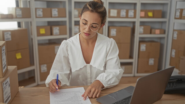 Woman holding document and pen, checking parcel labels and typing on laptop in storage building; small business concentration.