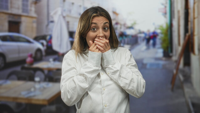Woman with hands covering mouth and fingers clasped, eyes closed, wearing white shirt on a restaurant terrace outdoors by sidewalk tables and umbrellas; surprise reaction.