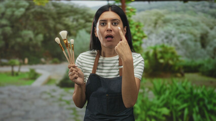 Fototapeta premium Woman wearing apron holding paintbrushes points finger to cheek on a forest path while holding makeup brushes and face cream; serious focus.