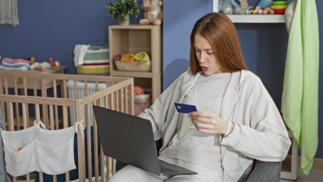 Woman redhead holding a creditcard and using a laptop while seated beside a baby crib and nursery storage shelves in a nursery; parenting multitasking contentment.
