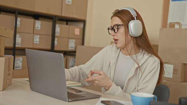 Redhead woman with headphones typing on laptop while gesturing with hand among stacked shipping boxes in building; small business focus.