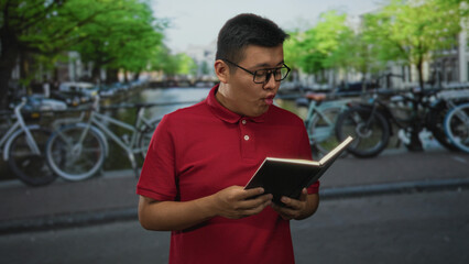 Obraz premium Man reading book on canal street beside bicycles and water, smiling while holding a notebook; urban leisure joy.