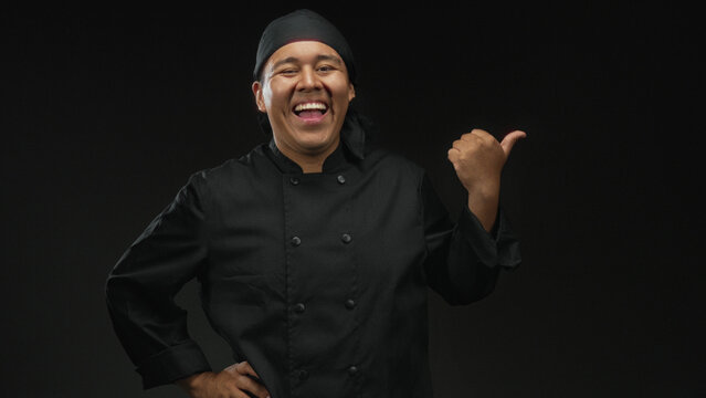 Man chef in black cook uniform pointing thumb to side with hand on hip in studio under black lighting; pride skill.