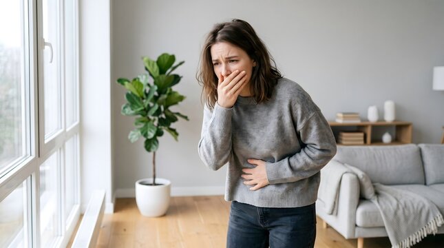 Distressed young woman suffering from acute nausea and stomach discomfort, possibly experiencing symptoms of illness, food poisoning, or morning sickness in her modern apartment
