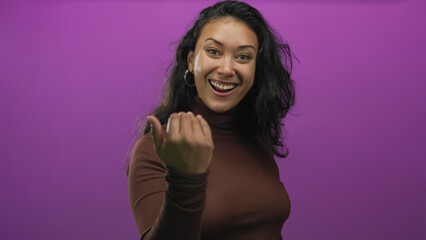 Woman pointing finger toward camera in a purple studio, smiling and beckoning while wearing hoop earrings and a brown turtleneck  playful invitation. © Krakenimages.com