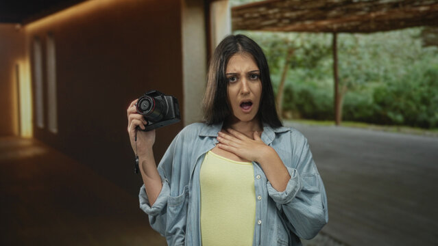 Young hispanic woman holding camera with hand on chest and mouth open in empty studio room; surprise.