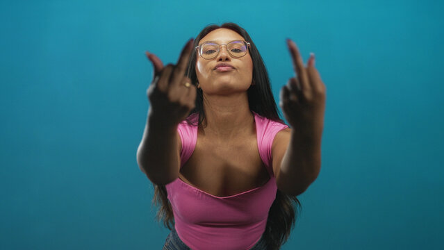 Woman showing both middle fingers with hands extended, wearing pink top and glasses in studio; defiance attitude.