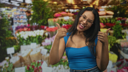 Fototapeta premium Woman in blue top showing thumbs up with visible thumbs and broad smile among bouquets at an indoor flower market building; joy confidence.