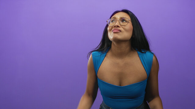 Woman covers ears with hands and winces in a purple studio backdrop wearing blue top and jeans; annoyance sensory overload.