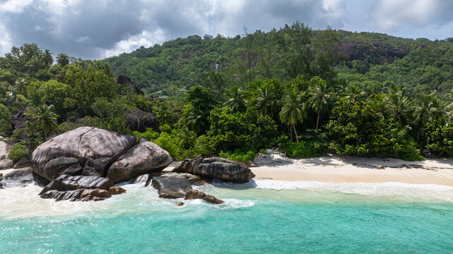 Granite boulders situated by turquoise ocean waters, set against lush forested hills. Seychelles, Mahe. Roche Copra.