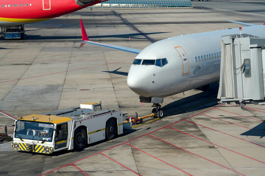 White passenger aircraft gets pushback from its terminal gate by an airport tug. A jet bridge connects to the plane. Another red airplane rests on the sunny tarmac