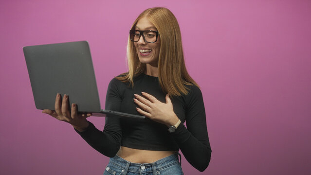 Woman holding laptop, smiling and waving hand with midriff visible in studio pink backdrop, wearing glasses and jeans; virtual meeting joy.
