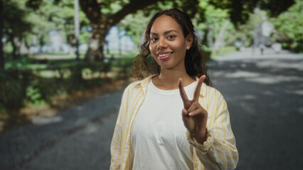 Fototapeta premium Woman wearing white tee and yellow striped shirt, palm up presenting on tree lined park path while touching hair; carefree confidence.