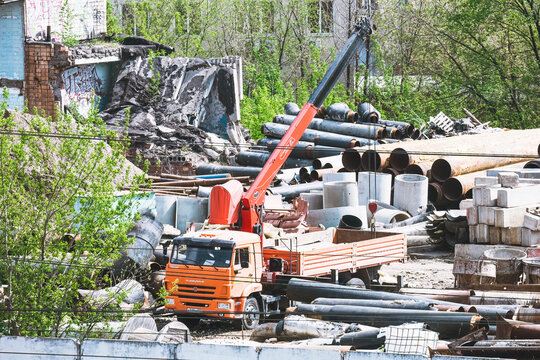 construction and demolition site. In foreground, there is an orange KAMAZ truck equipped with a boom crane, which is extended upwards. truck is parked among large assortment of construction materials