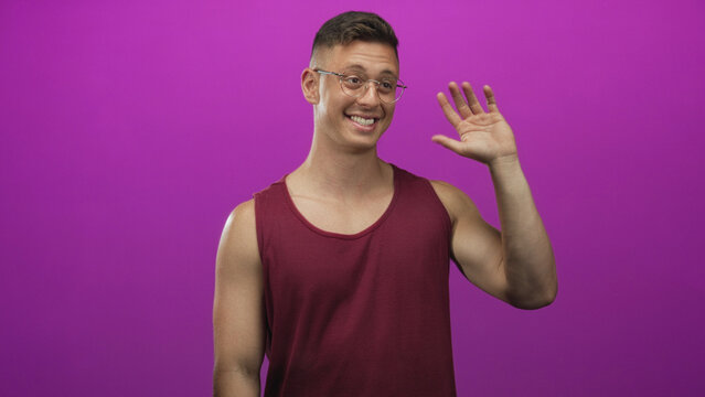 Young man in maroon tanktop and glasses points finger upward, bare arm visible, smiling in studio purple backdrop; greeting cheerful.
