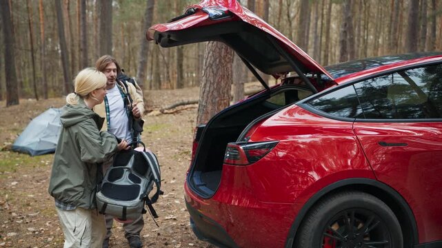 Caucasian couple carrying camping equipment near red car in forest. Blonde woman and handsome man unloading backpacks beside open trunk. Travelers preparing outdoor hiking adventure.