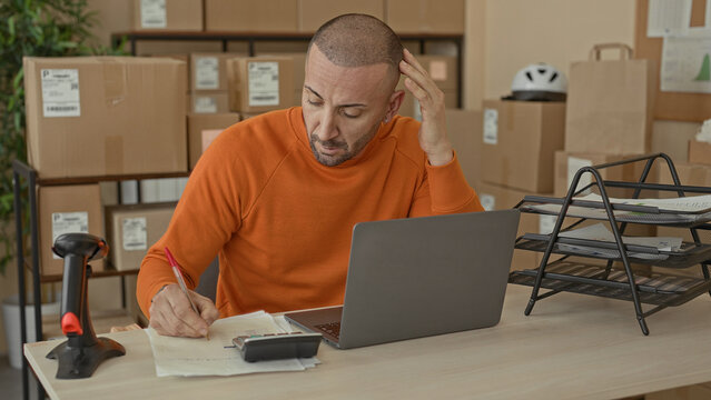 Man writing bills at a desk with laptop and cardboard packages, hand on head gesture in a parcel office building; small business stress.