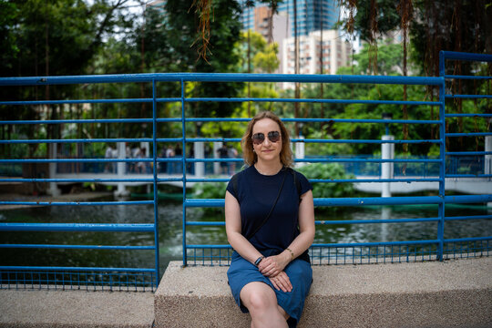 Woman sitting by a blue fence in a park with a pond and city buildings in background