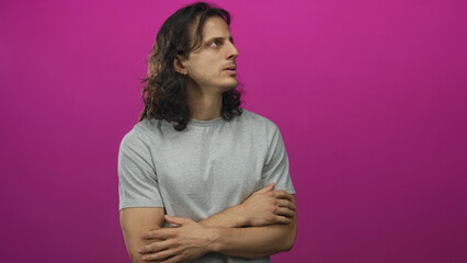 Obraz premium Young hispanic man with long hair and arms crossed in magenta studio wearing grey t shirt, looking sideways and contemplative; introspection.