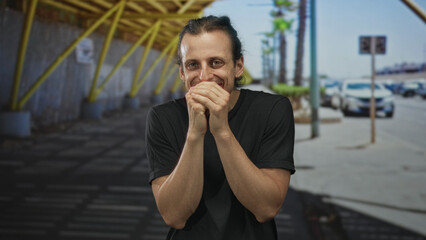 Fototapeta premium Man with hands over mouth smiling on street by parked cars and palm trees; nervousness anticipation.