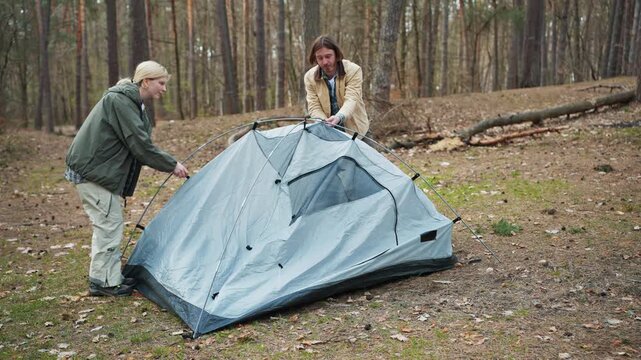 Blonde woman and Caucasian man assembling camping tent in forest. Couple preparing temporary shelter during hiking trip. Active travelers spending weekend outdoors in woodland area.