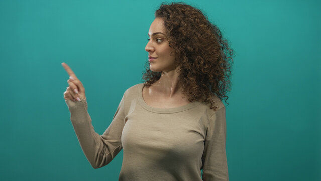 Young hispanic woman pointing finger with hand toward upper left while looking aside in studio with teal background; confidence focus.