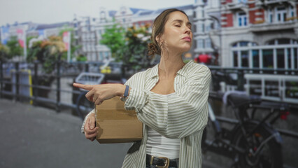 Fototapeta premium Woman holding cardboard box points finger on a city street canal bridge while carrying parcel in a delivery role; confident determination.