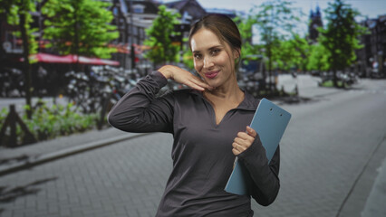 Fototapeta premium Woman holding clipboard and placing right hand under chin while smiling on street; playful confidence coaching.