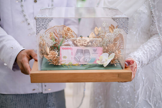 A couple holding a decorative glass box containing Indonesian currency as a traditional wedding dowry during a formal marriage ceremony.
