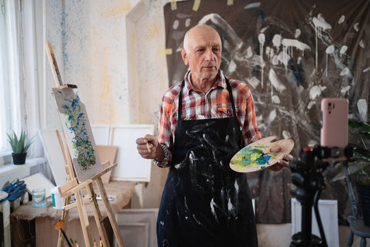 Senior elderly painter standing with drawing palette making video recording on smartphone using tripod in art creative studio.
