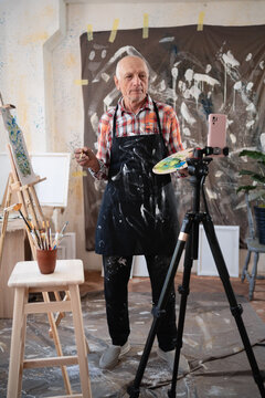 Senior elderly painter standing with drawing palette making video recording on smartphone using tripod in art creative studio.