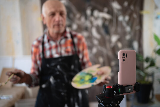 Senior elderly painter standing with drawing palette making video recording on smartphone using tripod in art creative studio.