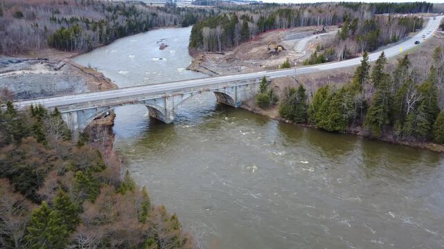 Drone video capturing a pickup truck with trailer and a car crossing the Arthur-Bergeron Bridge over the flooded Mitis River beside an active excavation site in spring 2026.
