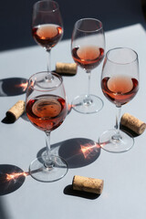 Obraz premium Four glasses filled with rose wine sit on grey surface. Direct sun creates shadows and light patterns. Wine corks rest near glasses. Simple composition.