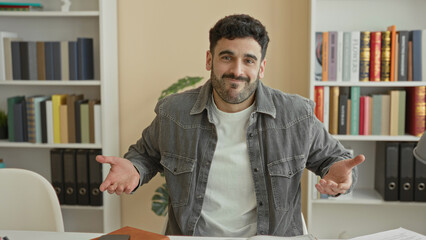Fototapeta premium Man shrugging with palms up at desk surrounded by books, papers and notebook in a studio workspace; puzzled uncertainty.