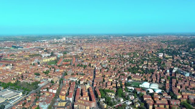 4K aerial view of Bologna residential neighbourhoods with dense terracotta rooftops, suburban streets, sports facilities and Po Valley plain stretching to the horizon in Italy.