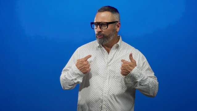 Man middle aged hispanic with grey beard and glasses pointing thumbs to chest in blue studio wearing patterned white shirt; self confidence.