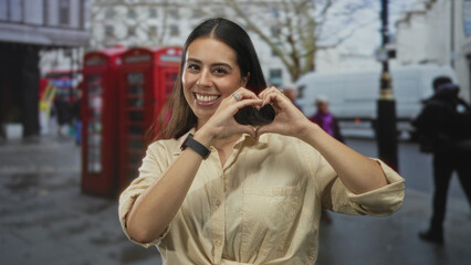 Fototapeta premium Woman forming a heart with hands on a city street by red telephone booths, smiling in beige shirt and showing rings on fingers; joy love.